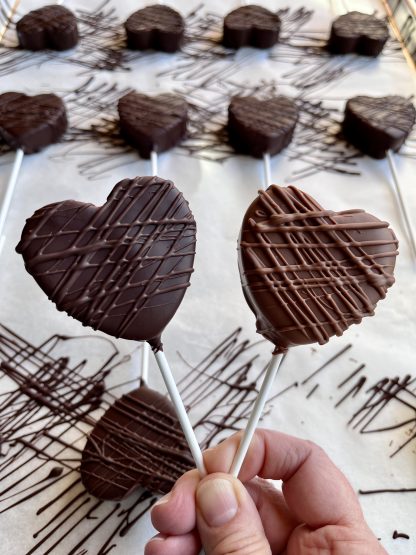 A hand holds two marshmallow heart lollipops. The marshmallow hearts are coated in Belgian chocolate. The background is a pan of more lollipops.