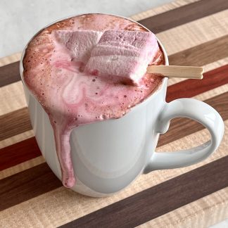 A mug of hot cocoa sits on a wooden board. There are two peppermint marshmallows in the hot cocoa. The cocoa and melted marshmallow has dripped over the edge of the mug.