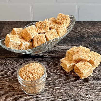 A clear glass dish filled with toasted coconut marshmallows sits on a brown board. There is also a small pile of the marshmallows on the board as well as a small jar filled with toasted coconut flakes. The background is white subway tile.