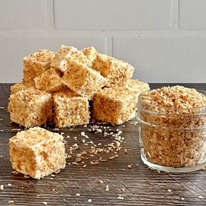 A pile of toasted coconut marshmallows sits on a brown board. Toasted coconut flakes are scattered on the board. There is also a small jar filled with toasted coconut flakes. The background is white subway tile.