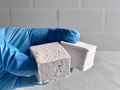 A hand wearing a blue vinyl glove holds two lavender honey marshmallows over a white marble board. The background is white subway tile.