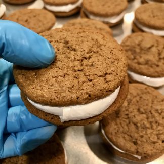A gloved hand holds an oatmeal creme pie over a tray of more oatmeal creme pies.