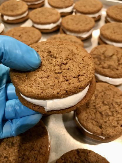 A gloved hand holds an oatmeal creme pie over a tray of more oatmeal creme pies.