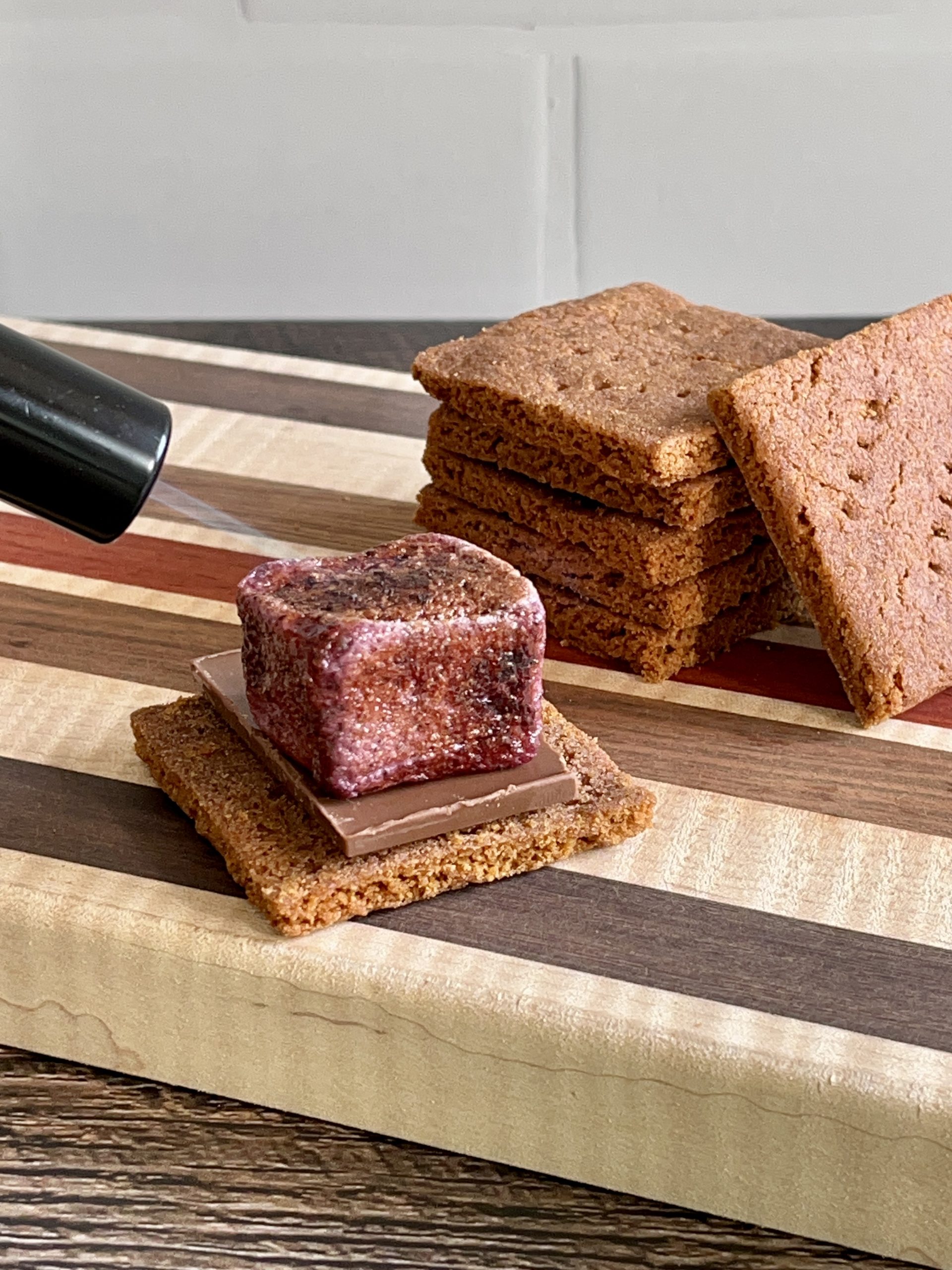 A small kitchen torch is being used to toast a wild blueberry marshmallow. The marshmallow is on top of a graham cracker with a piece of chocolate. There is a stack of graham crackers in the background.