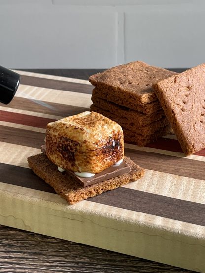 A small kitchen torch is being used to toast a vanilla marshmallow. The marshmallow is on top of a graham cracker with a piece of chocolate. There is a stack of graham crackers in the background.