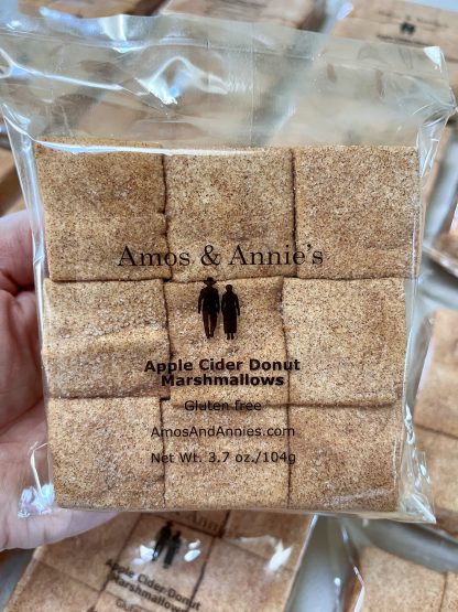 A hand holds a package of apple cider donut marshmallows above other packages on a table.